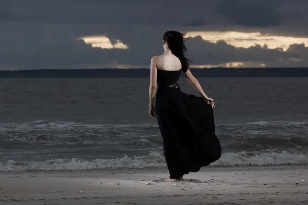 Vacker brunett modell i lång svart klänning stående på stranden. molnigt väder. Sunset — Stockfoto