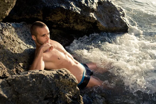Fitness chlap ležící na kámen ve vodě. krátké vlasy. Butch. černé plavky. opálenou pleť. ruce za hlavu. nasměrovat. západ slunce — Stock fotografie
