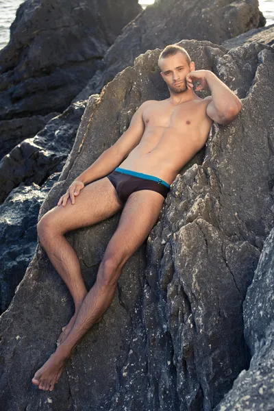 Ragazzo con il corpo perfetto sdraiato sulla pietra. capelli corti. Butch. costumi da bagno neri. pelle abbronzata. piena crescita — Foto Stock