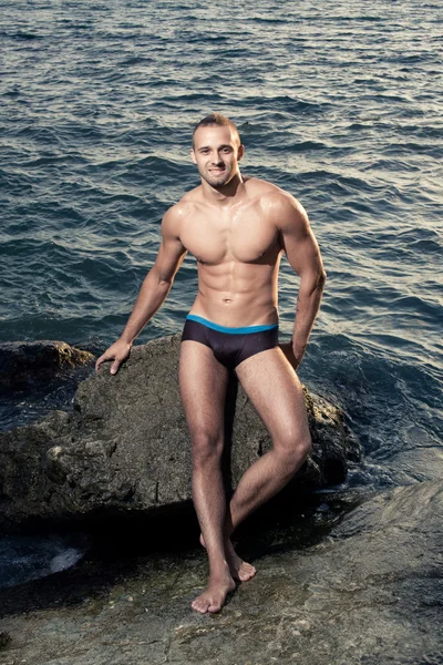 Bodybuilder sorridente in riva al mare. piena crescita. Capelli corti. Butch. costumi da bagno neri. pelle abbronzata . — Foto Stock