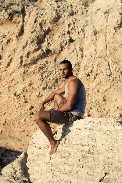 Hombre de cuerpo perfecto sentado en la orilla del mar sobre fondo claro. camiseta gris, pantalones marrones — Foto de Stock