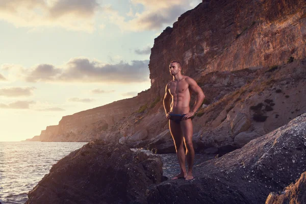 Perfect body guy posing on seascape and mountainscape background.black trunks, black swimsuit. short hair. serious look. HDR. evening. sunset