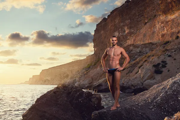 Bodybuilder-Mann posiert vor Meeres- und Bergkulisse. Abend. Sonnenuntergang.Schwarze Badehose, schwarzer Badeanzug. kurze Haare. Ernster Blick. hdr — Stockfoto