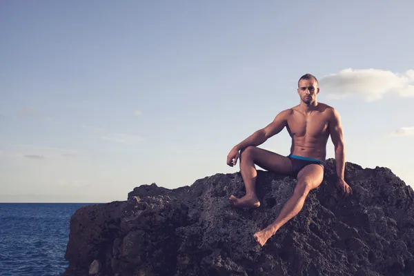 Typ mit perplexem Körper, der auf Felsen sitzt. schwarze Badehose, schwarzer Badeanzug. kurze Haare. Ernsthafter Blick — Stockfoto