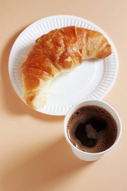 Kağıt tabakta kruvasan, bej arka planda kahve fincanı, üst manzara paket yemek.