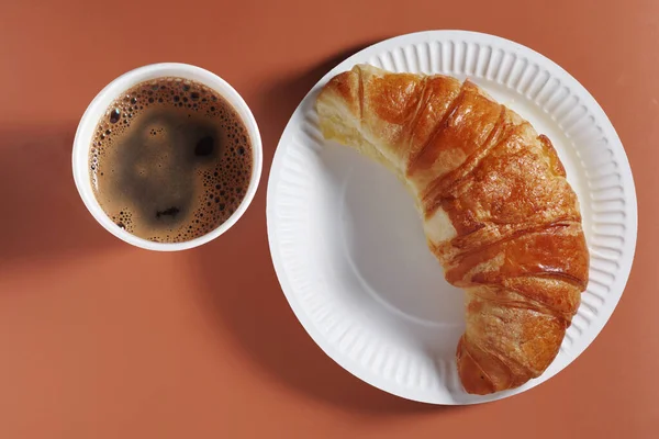 Croissant on paper plate and coffee cup on brown background, top view Takeaway food