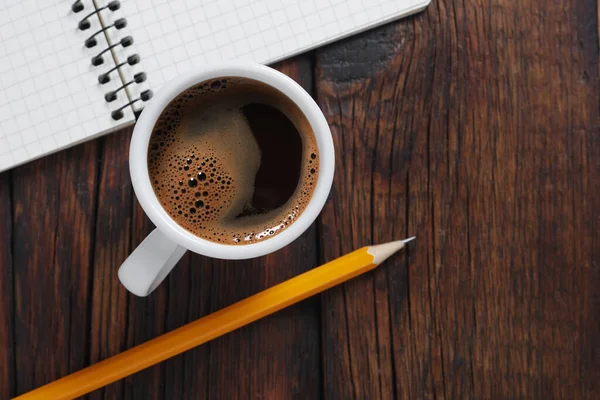 Cup of coffee and spiral notepad with pencil on wooden table close up, top view