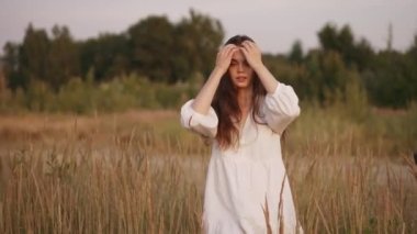Girl in white dress walks around the field, touches her hair and looks into the camera.