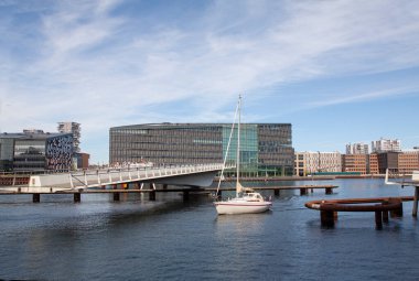 Sailing yacht passing through an open modern moveable pedestrian swing bridge in urban harbor with buildings and blue sky in the background. Copy space.
