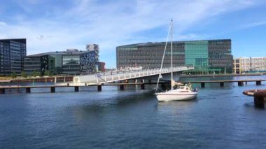 Şehir limanındaki hareketli bir yaya köprüsünden geçen yelkenli bir yat. Arka planda binalar ve mavi gökyüzü var.. 
