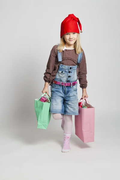 微笑女孩携带圣诞购物袋女の子身に着けているクリスマスの帽子に笑みを浮かべて、つまらないものを保持しています。 — 图库照片