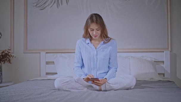 Feminino vestindo pijama digitando mensagem no celular no quarto — Vídeo de Stock