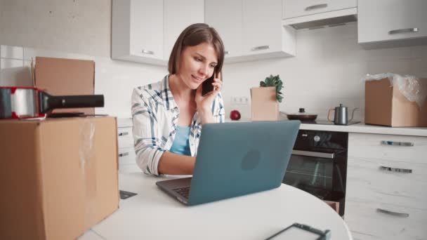 Mujer hablando por teléfono escribiendo en el ordenador en la cocina — Vídeos de Stock