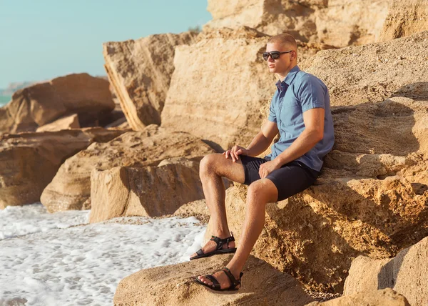 Fashionable young muscular guy resting on a rocky beach. — Stock Photo, Image