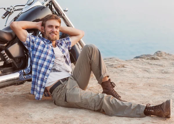 Homem está sentado na praia com as costas para a moto . — Fotografia de Stock