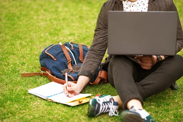 นักเรียนหนุ่มแฟชั่นชายนั่งบนหญ้าในสวนสาธารณะและถือ — ภาพถ่ายสต็อก