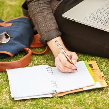 young male student sitting on grass in park and holding a laptop clipart