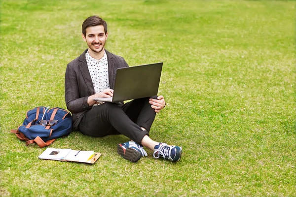 Молодой человек использует свой ноутбук на траве и делает заметки в не — стоковое фото