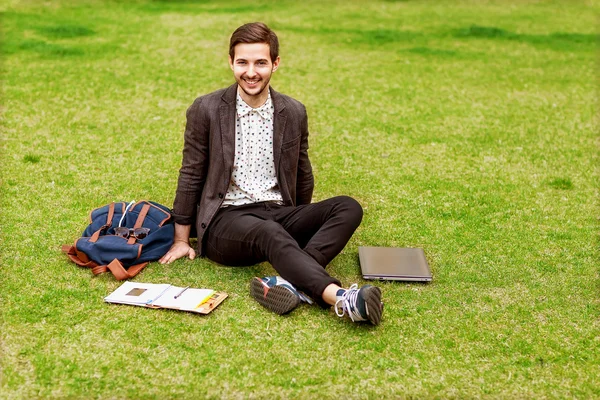 Jovem moda estudante do sexo masculino sentado na grama no parque e segurando — Fotografia de Stock