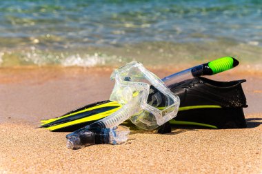 Kumda yüzen ekipmanlar ve okyanus dalgaları su sıçratıyor. Siyah yüzgeçler, siyah maske, kumlu dokuda şnorkel. Kumda yatan eşyalar.
