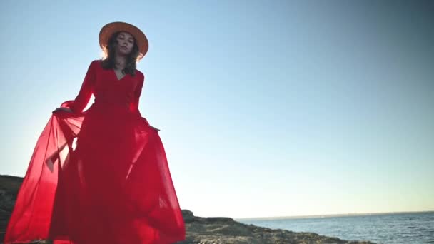 Slow motion young caucasian woman in a loose fluttering red dress is walking along the rocky seashore. Shallow depth of field. Offset focus. — Stock Video