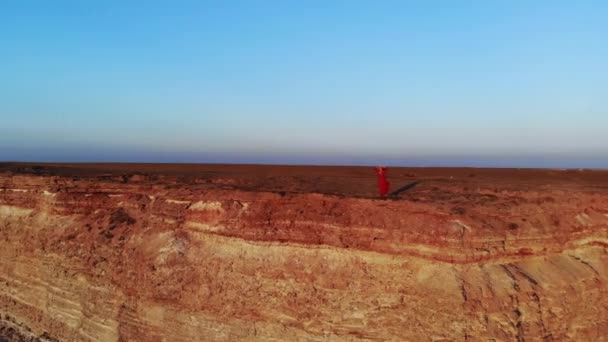Eine attraktive junge Frau in rotem Kleid und Strohhut steht bei windigem Wetter am Rande einer Klippe an einer hohen Felsküste. Luftaufnahme — Stockvideo