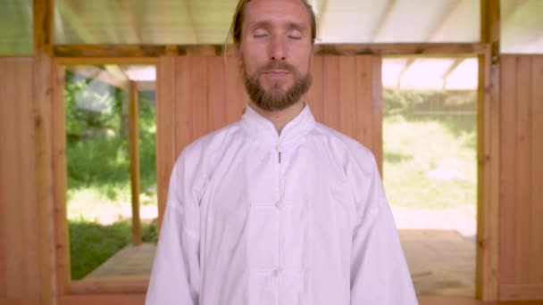 Ein bärtiger Kaukasier in heller Kleidung praktiziert Qigong im hölzernen Raum der Halle. Moderne Traditionen der Energiepraxis — Stockvideo
