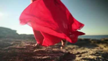 Gevşek kırmızı elbiseli, ağır çekim beyaz kadın kayalık deniz kıyısında yürüyor. Yüzeyin derinliği. Yerleşim odağı.