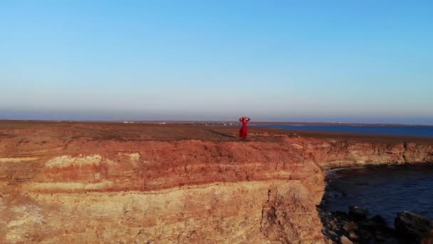 Приваблива молода жінка в червоній сукні і солом'яний капелюх у вітряну погоду стоїть на краю скелі високого кам'янистого узбережжя. Вид з повітря — стокове відео