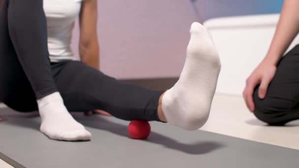 Close-up Jovem massagista do sexo masculino ensina cliente mulher caucasiana paciente miofascial auto-massagem de pernas e quadris usando bola de massagem dentro de casa. Libertação do miofasceal — Vídeo de Stock