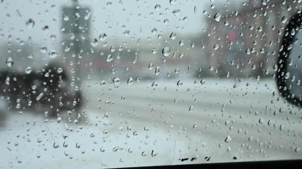 Капли растаявшего снега на боковое стекло автомобиля. Не в фокусе заснеженные городские улицы с автомобильным движением — стоковое видео