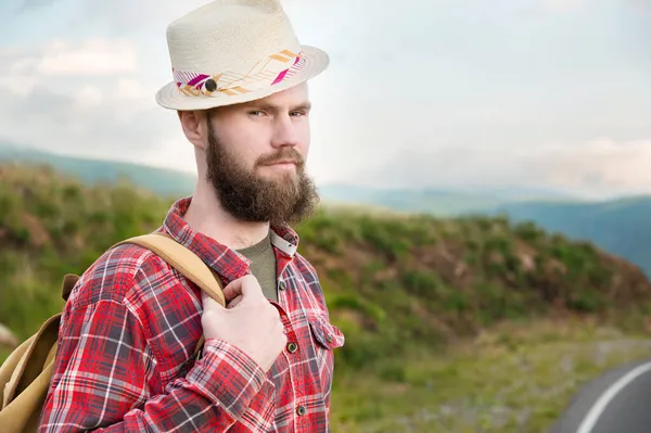 Homem caucasiano barbudo romântico atraente usando um chapéu, camisa xadrez e uma mochila. Fica no campo nas montanhas e olha para a câmera. Espaço para cópia. Caroneiro — Fotografia de Stock