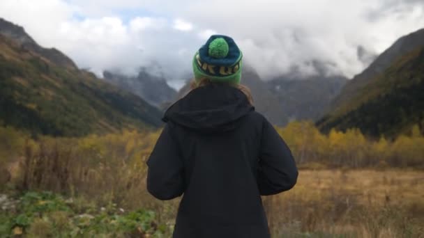 Ondiepe scherptediepte, focus verschuiving. Een jonge onherkenbare vrouw in een jasje en wintermuts staat in een bergkloof en kijkt naar de bergen. Zicht van achteren. Groot dynamisch bereik — Stockvideo