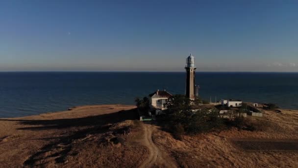 Vista aérea de un faro solitario rodeado de edificios técnicos se encuentra en un promontorio rocoso desierto en la orilla del mar. Oceánica costa de la tarde. Faro al atardecer. Bajo perfil. grano de película — Vídeo de stock