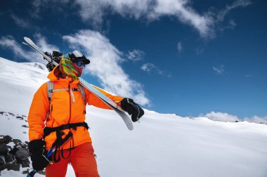 Omuzlarında kayak sopaları ve kayaklarla turuncu giyimli profesyonel bir kayakçının dağ portresi. Karlı bir yamaçta, yüksek dağlarda