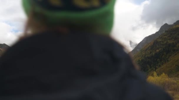 Крупный план Небольшая глубина резкости, смещение фокуса. Молодая неузнаваемая женщина в пиджаке и зимней шляпе стоит в альпийском ущелье и смотрит на горы. Вид со спины. Высокая динамика — стоковое видео