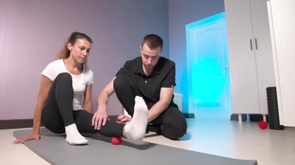 Wypuszczenie na rynek. Męski fizjoterapeuta umieszcza piłkę do rehabilitacji mięśni nóg klientów siedząc na macie do jogi, masując technikę pacjenta czerwoną piłką. — Wideo stockowe