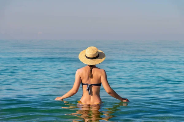 İnce, beyaz bir kadının, beline kadar durgun, şeffaf deniz suyuna batmış, mayosu ve hasır şapkasıyla duruşu.