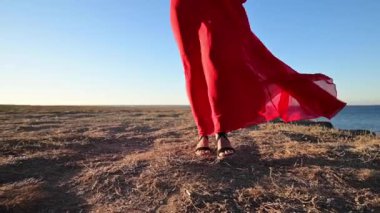 Ağır çekim. Kırmızılı, gevşek elbiseli ve sandaletli beyaz bir kadının ince bacakları kayalık yüksek deniz kıyısında geziniyor.