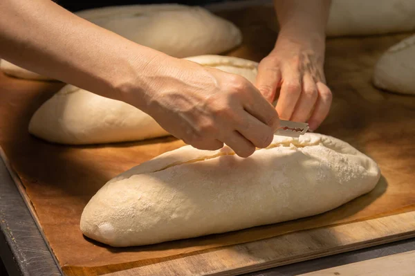 Primer plano de un panadero manos femeninas cortar una hogaza de masa en una hogaza de pan con una hoja antes de hornear en el horno. Producción artesanal de pan artesanal —  Fotos de Stock