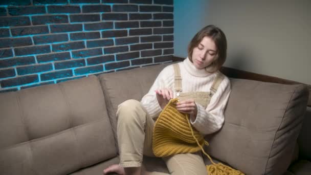 Μια νεαρή λευκή ελκυστική γυναίκα με καθημερινά ρούχα κάθεται στον καναπέ στο δωμάτιο και πλέκει ένα καπέλο από μαλλί. Χόμπι και κατασκευή ειδών ένδυσης στο σπίτι — Αρχείο Βίντεο
