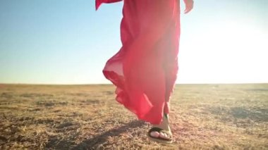 Kırmızı elbiseli bir kadının kuru çimenlerde yürümesi sinematik ağır çekimde. Sandaletlerle kumlu bozkırın dalgalı yüzeyinde rüzgarda sallanan elbiseli bir kadın gezgin. 4K Sahne Kadın Doğası.