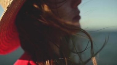 Rüzgarda yavaş çekimde dalgalanan Kafkasyalı genç bir kadının yakın çekimi. Yüzeyin derinliği. Kırmızı elbiseli ve şapkalı kız deniz kenarında yürüyor.