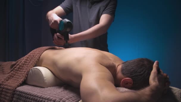 Beyaz profesyonel erkek masör, perküsyon vibro masaj aleti kullanarak sırt ve sırt kasları için masaj yapıyor. Masajı yenilemek için şok tedavisi — Stok video