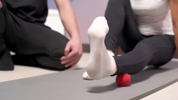 Миофасциальное освобождение крупным планом. Мужчина физиотерапевт помещает мяч для реабилитации мышц ног клиента, сидя на коврике для йоги, массируя технику пациентов с помощью красного шара. — стоковое видео