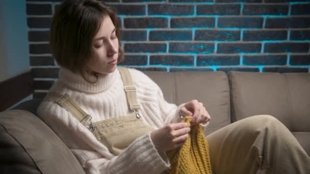 Jovem mulher caucasiana atraente em roupas casuais tricô um chapéu de lã enquanto crochê enquanto sentado no sofá em casa. Hobbies de artesanato e fabricação de roupas para casa — Vídeo de Stock