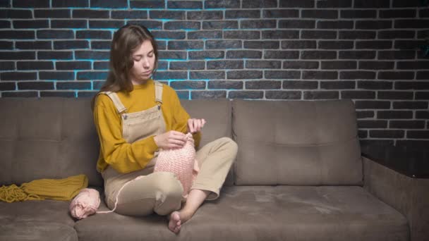 Attractive Caucasian young woman in casual clothes knitting a wool hat while crocheting while sitting on the sofa at home. Handicraft hobbies and home clothing making — Stock Video