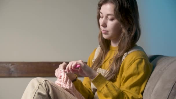 Attractive Caucasian young woman in casual clothes knitting a wool hat while crocheting while sitting on the sofa at home. Handicraft hobbies and home clothing making — 图库视频影像