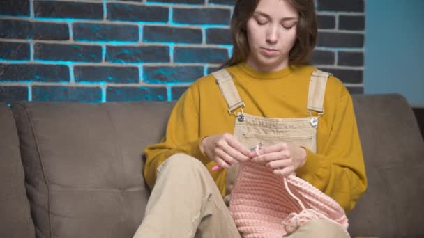 Молодая кавказская привлекательная женщина в повседневной домашней одежде сидит на диване в комнате и вяжет шляпу из шерсти. Хобби и домашняя одежда — стоковое видео