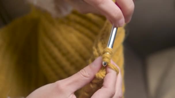 Close-up. Hands of an attractive young caucasian woman in casual clothes sitting on a sofa indoors knits a woolen product by a crochet — Stock Video